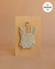 A Mrs. Ertha Doll Clothes - 13″-14″ - Little Daisys Frilled Bodysuit, featuring delicate white floral patterns and frilled details in beige, hangs on a small hanger against a wooden board. Ideal for dressing Minikane dolls, it includes a label stating “fits MINKANE 13”.