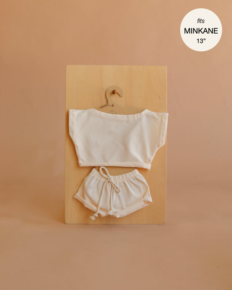 Introducing the Mrs. Ertha Doll Clothes - 13″-14″ - Ivory Comfy Set: This charming ivory outfit is crafted from organic cotton and includes a short-sleeve top and drawstring shorts, all displayed on a hanger against a wooden board. The corner text notes “fits Minikane 13’’.” Perfect as handmade doll clothes for your little one’s collection!.