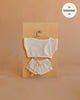Introducing the Mrs. Ertha Doll Clothes - 13″-14″ - Ivory Comfy Set: This charming ivory outfit is crafted from organic cotton and includes a short-sleeve top and drawstring shorts, all displayed on a hanger against a wooden board. The corner text notes “fits Minikane 13’’.” Perfect as handmade doll clothes for your little one’s collection!.