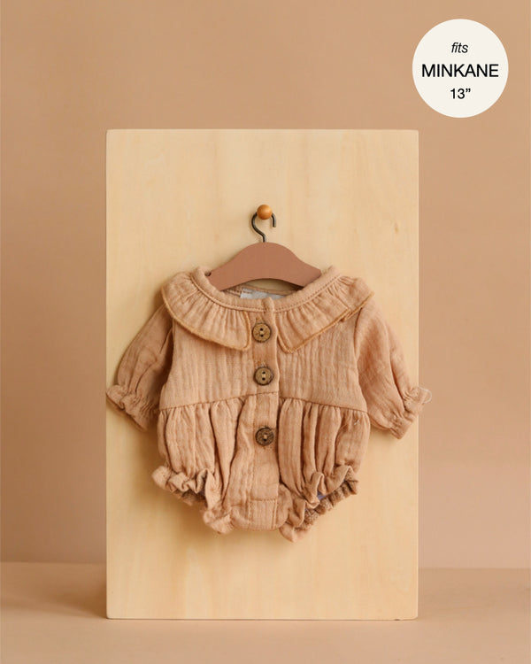 The Leonore Ruffled Romper in Mocha cotton gauze for Minikane dolls is shown on a small hanger against a wooden board, featuring long sleeves, ruffles, and large buttons. A label in the corner reads “fits MINIKANE 13”.