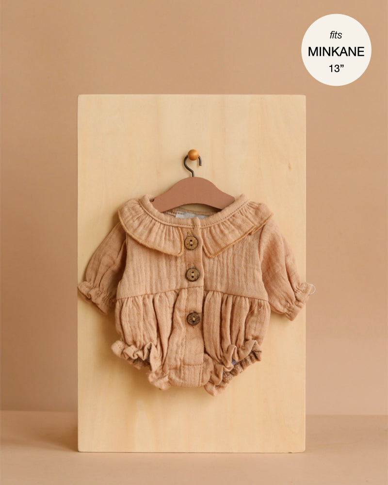 The Leonore Ruffled Romper in Mocha cotton gauze for Minikane dolls is shown on a small hanger against a wooden board, featuring long sleeves, ruffles, and large buttons. A label in the corner reads “fits MINIKANE 13”.