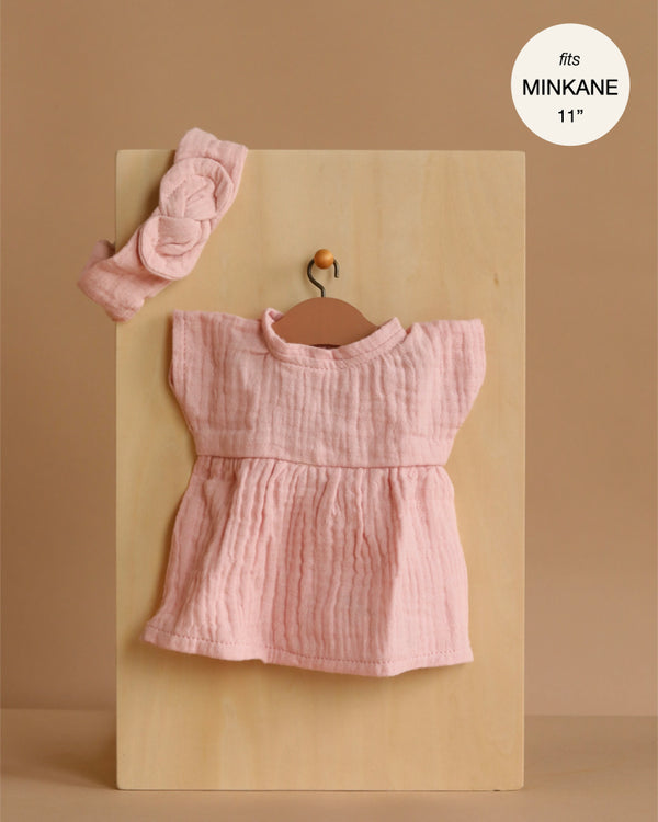 The Minikane 11" Doll Clothing set features the Faustine Dress & matching headband in cotton gauze, Petal Pink, displayed on a wooden hanger against a beige background. A label notes it fits Minikane Babies dolls 11”.