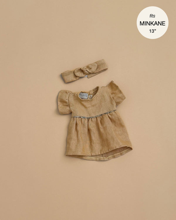 The Minikane Doll Clothing Daisy Dress & Headband in Washed Linen - Oatmeal features a small beige dress with puffed sleeves and gathered waist, plus a matching bow headband, displayed on beige. Fits Minikane and 13-inch dolls.