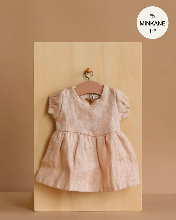 The Faustine Dress with Balloon Sleeves in Nude Linen, made for Minikane 11" dolls, hangs on a wooden hanger against a light wood board and beige background. A white circle notes "fits Minikane Babies dolls 11”".