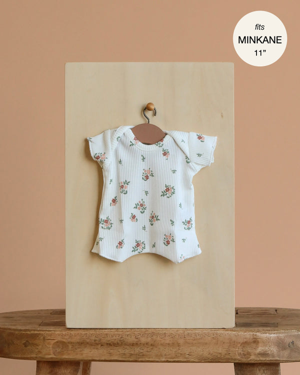 The Minikane 11" Doll Clothing | Shorty Bodysuit in Ribbed Knit - Small Flowers, a white ribbed cotton short-sleeve romper with a pink floral print, is displayed on a hanger with a label reading "fits MINIKANE 11''.