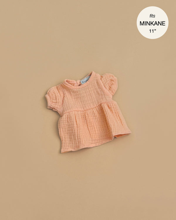 Apricot cotton gauze Faustine dress for Minikane 11" dolls, featuring short balloon sleeves, shown laid flat on a beige background. Text in the top right corner reads “fits MINIKANE 11”.”.