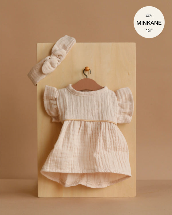The Minikane 13" Doll Clothing set features a putty-colored daisy dress with flutter sleeves and a matching knotted headband in cotton gauze. Displayed on a wooden hanger, this set fits Minikane 13" dolls. Text: "fits MINKANE 13".