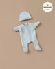 A cream-colored ribbed onesie with long sleeves and footed legs, paired with a matching beanie, rests on a light brown background. Designed in France for Minikane dolls, the circular label reads "fits MINKANE 11''". Product: Minikane Doll Clothing | Sleep well Angel and her linen ribbed hat.