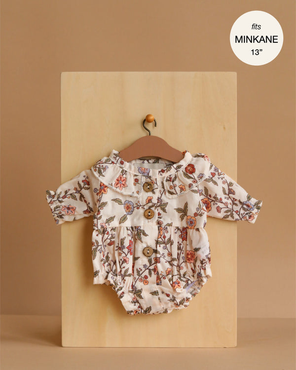 The Minikane 13" Doll Clothing | Leonore Ruffled Romper in Cotton Gauze - Gisele, featuring floral patterns, long sleeves, and wooden buttons, is displayed on a wooden hanger against a beige background. "Fits MINIKANE 13"" appears in the top right corner.