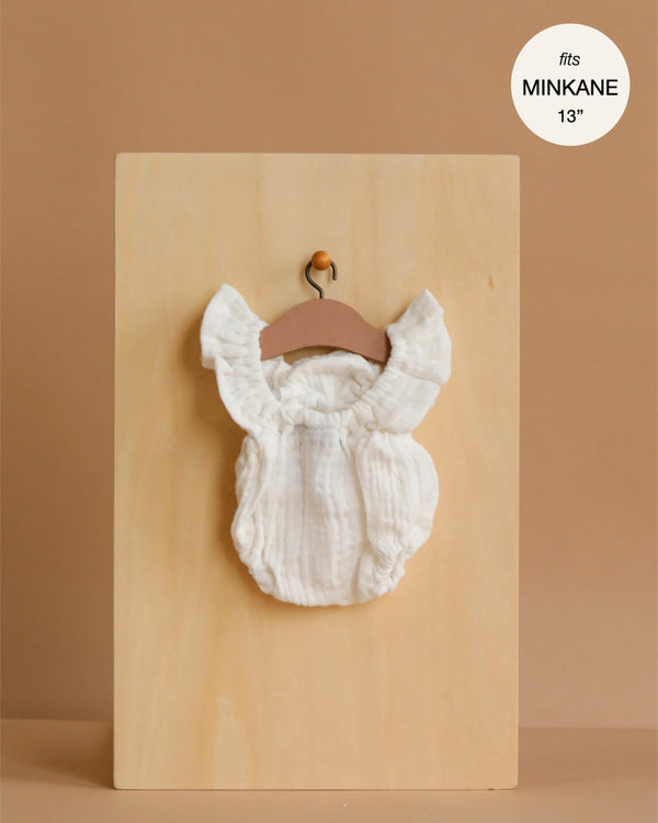 A cream, ruffled Lou Retro Romper in cotton gauze for Minikane 13" dolls hangs on a wooden hanger against a plain wooden board background.