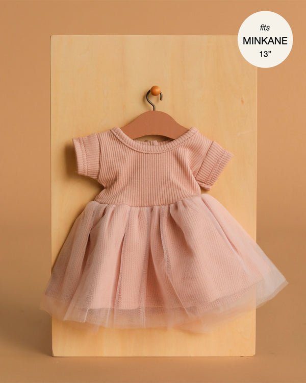 The Minikane Doll Clothing | Rosana Short Sleeve Tutu in Petal Pink features a ribbed knit bodice and tulle skirt, sized for 13" Minikane dolls. Displayed on a wooden hanger against wood panels. Made in France—perfect 13 inch doll clothes.