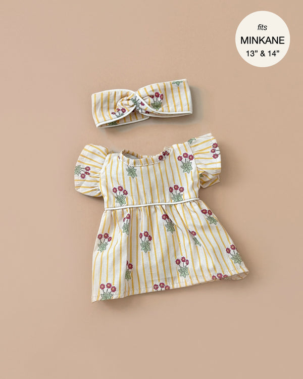 The Daisy Dress and matching crossed headband, designed for Minikane dolls in a yellow and white striped floral pattern with puffed sleeves and an empire waist, are hand washable. Crafted in France from cotton daisies with piping.