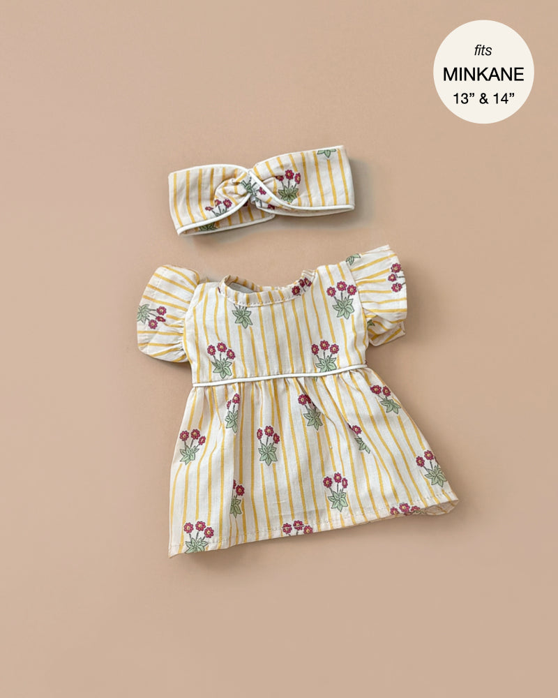 The Daisy Dress and matching crossed headband, designed for Minikane dolls in a yellow and white striped floral pattern with puffed sleeves and an empire waist, are hand washable. Crafted in France from cotton daisies with piping.