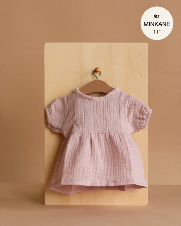 An orchid pink, balloon-sleeved Faustine dress in cotton gauze for 11" Minikane dolls hangs on a small wooden hanger against a neutral beige wooden board. Designed in France.
