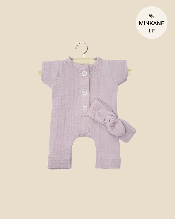 The Lili jumpsuit in lavender, ideal for Minikane Gordis dolls, features short sleeves, white buttons, and a fabric bow. Crafted in France with a label "fits MINKANE 11", it includes a petal cotton gauze headband and is displayed on a hanger against a soft beige backdrop.