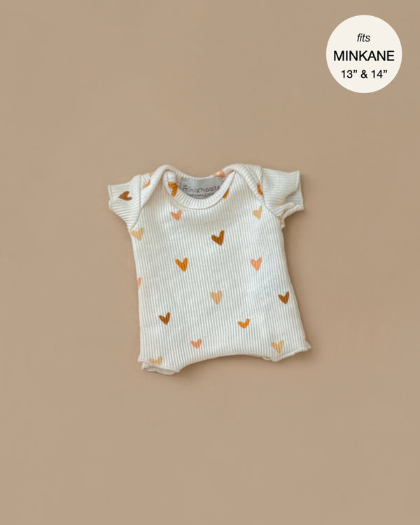 The Minikane Doll Clothing Les P'tits Basique Ribbed Bodysuit in Hearts is a beige, cream-colored short-sleeve onesie made from soft cotton with golden and brown heart patterns for Minikane 13" & 14" dolls. Designed in France, it fits Gordis dolls.