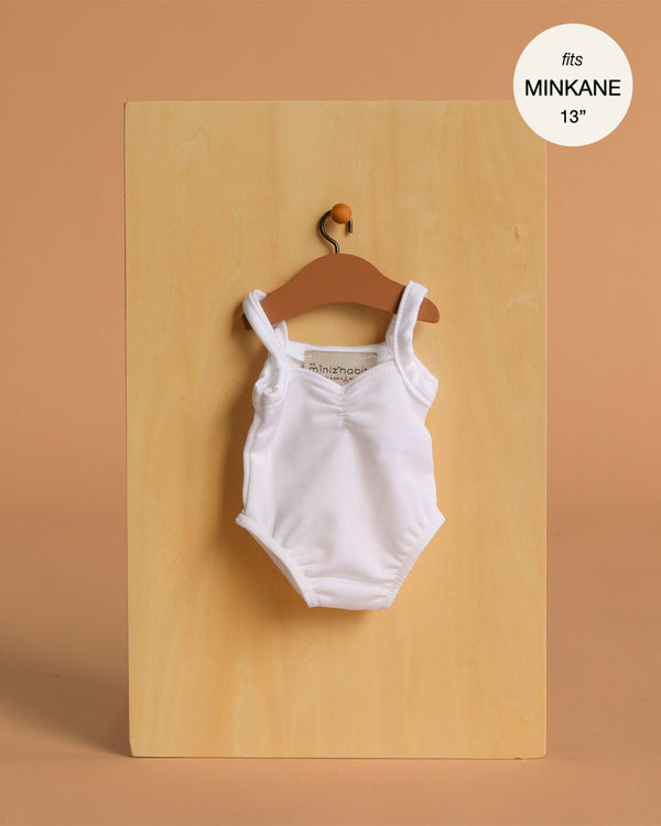 A white Anna Leotard in shiny lycra for Minikane 13" dolls hangs on a brown hanger against a light wooden board.