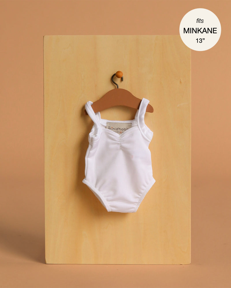 A white Anna Leotard in shiny lycra for Minikane 13" dolls hangs on a brown hanger against a light wooden board.