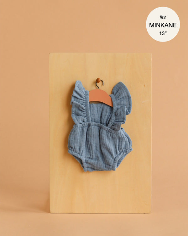 The Lou Retro Romper in blue denim cotton gauze for Minikane 13" dolls is shown on a wooden hanger against a beige background. A round label reads “fits MINKANE 13”.” Ideal for any 13-inch doll clothing collection.