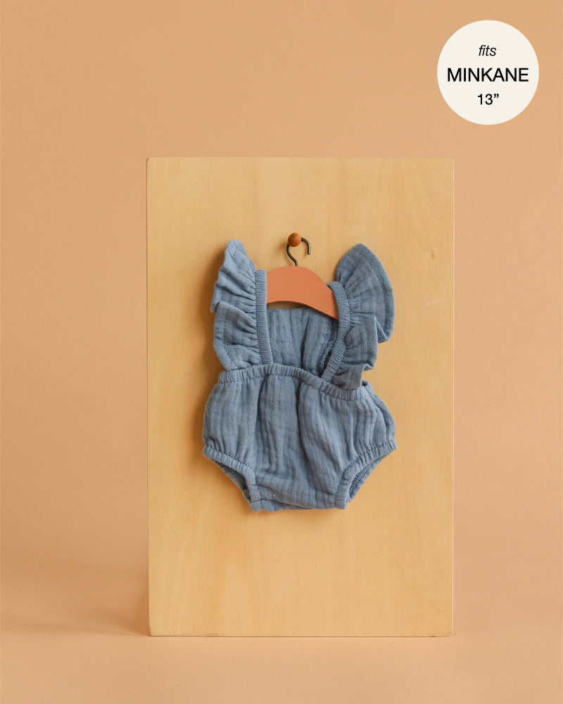 The Lou Retro Romper in blue denim cotton gauze for Minikane 13" dolls is shown on a wooden hanger against a beige background. A round label reads “fits MINKANE 13”.” Ideal for any 13-inch doll clothing collection.
