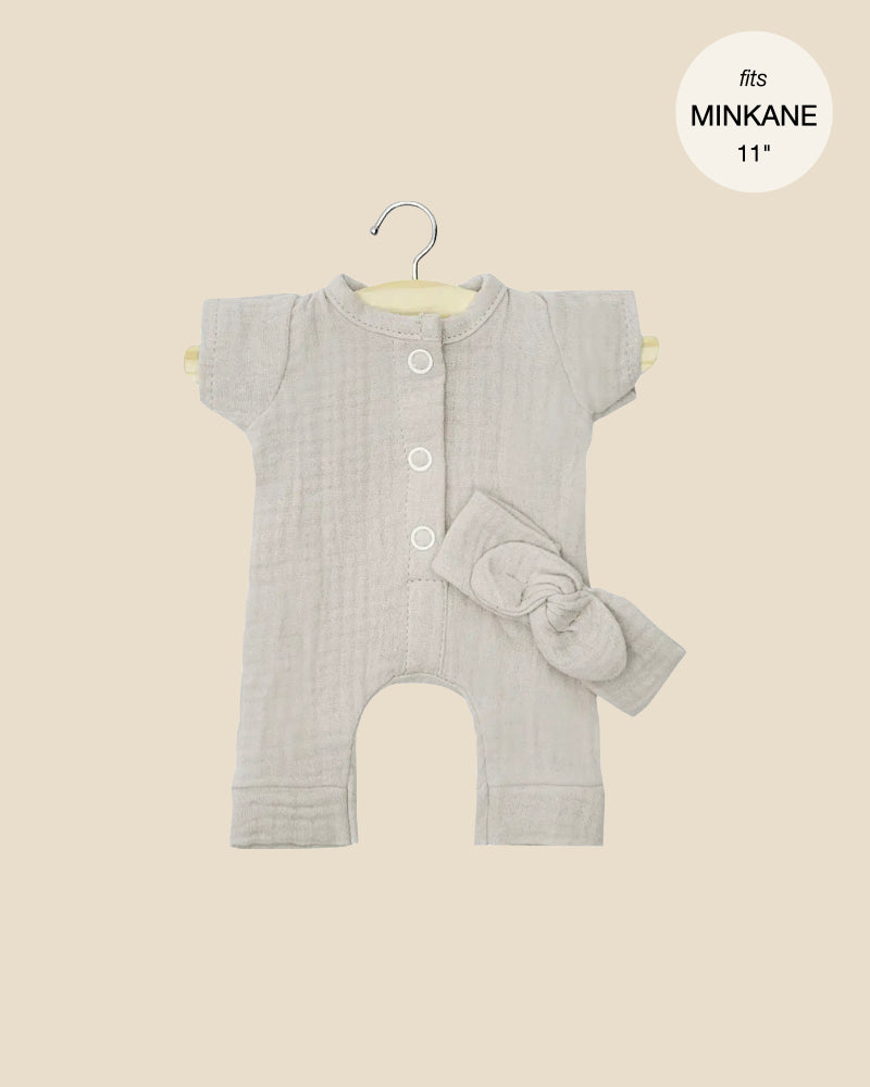 The Minikane Doll Clothing set includes a Lili Jumpsuit with short sleeves and four white buttons, perfect for 11" dolls. Displayed on a hanger against a beige backdrop, it comes with a pebble cotton gauze headband bow and ships in approximately one week.