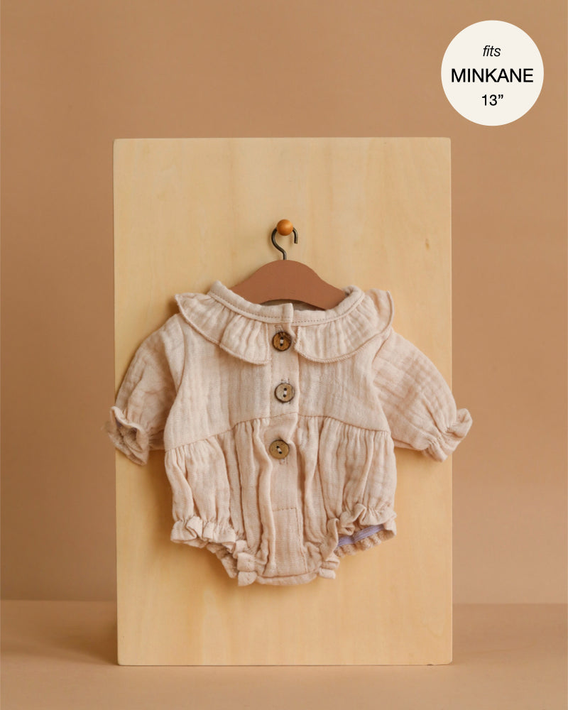 The Minikane 13" Doll Clothing Leonore Ruffled Romper in Putty cotton gauze features long sleeves, a ruffled collar, and wooden buttons. Displayed on a small hanger, it’s designed to fit 13-inch Minikane dolls.