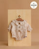 The Minikane 13" Doll Clothing Leonore Ruffled Romper in Putty cotton gauze features long sleeves, a ruffled collar, and wooden buttons. Displayed on a small hanger, it’s designed to fit 13-inch Minikane dolls.