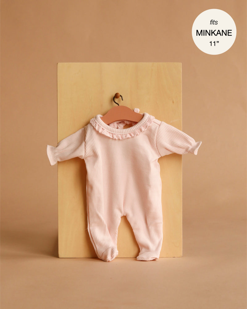 The Minikane 11" Doll Clothing | Camille Onesie in Ribbed Knit with Lace - Pastel Pink is a light pink, long-sleeved romper with lace collar detail for 11" Minikane Dolls, made in France and displayed on a wooden hanger against a tan background.