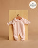 The Minikane 11" Doll Clothing | Camille Onesie in Ribbed Knit with Lace - Pastel Pink is a light pink, long-sleeved romper with lace collar detail for 11" Minikane Dolls, made in France and displayed on a wooden hanger against a tan background.