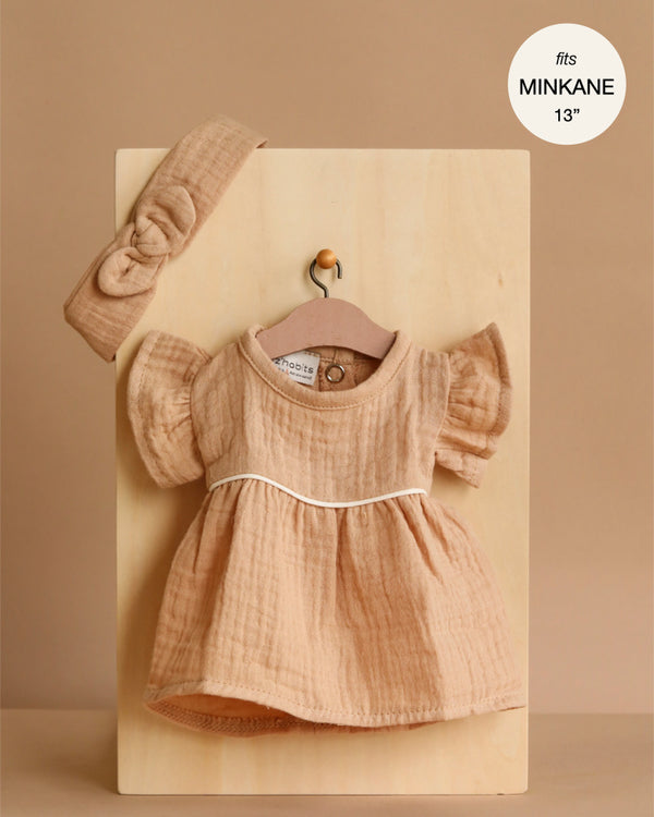 The Minikane Doll Clothing | Daisy Dress & Headband in Cotton Gauze - Latte is a light brown, short-sleeved dress with white trim, shown on a wooden board with a matching headband. Fits 13" Minikane dolls and can be styled for 14" dolls.