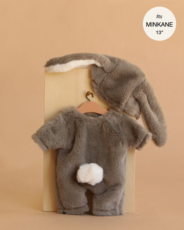 The Minikane 13" Doll Clothing | Bunny Costume includes a fuzzy gray short-sleeved suit with a white tail and a hat with floppy ears, shown on a hanger against a beige background. Designed to perfectly fit Minikane Gordis 13" dolls.