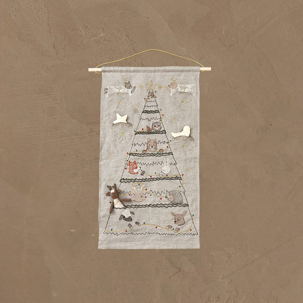 The Coral & Tusk Embroidered Advent Calendar hangs from a wooden rod and string on a plain brown textured wall, featuring a Christmas tree outlined and decorated with embroidered animals, stars, and ornaments.