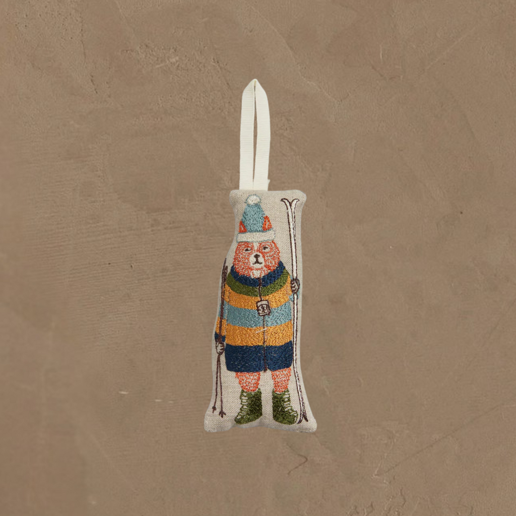 The Coral & Tusk Slopes Fox Ornament features a fox skier in a striped sweater, blue pants, green boots, and pom-pom cap, holding skis and poles. It hangs from a white loop against a beige textured background.