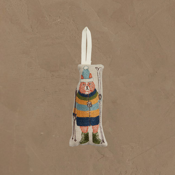 The Coral & Tusk Slopes Fox Ornament features a fox skier in a striped sweater, blue pants, green boots, and pom-pom cap, holding skis and poles. It hangs from a white loop against a beige textured background.