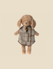 A Maileg Puppy Poncho with brown fur is wearing a checkered coat with a small tail hole. The light-colored coat, adorned with brown stripes, features a small pocket on the front. The dog stands against a beige background.