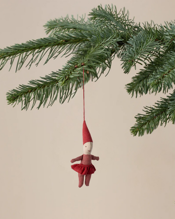 The Maileg Pixy Ornament - Girl, a small fabric doll with a red hat and striped outfit, hangs from a green pine branch, bringing festive charm to your Christmas decor against a neutral beige background.