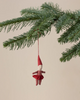The Maileg Pixy Ornament - Girl, a small fabric doll with a red hat and striped outfit, hangs from a green pine branch, bringing festive charm to your Christmas decor against a neutral beige background.