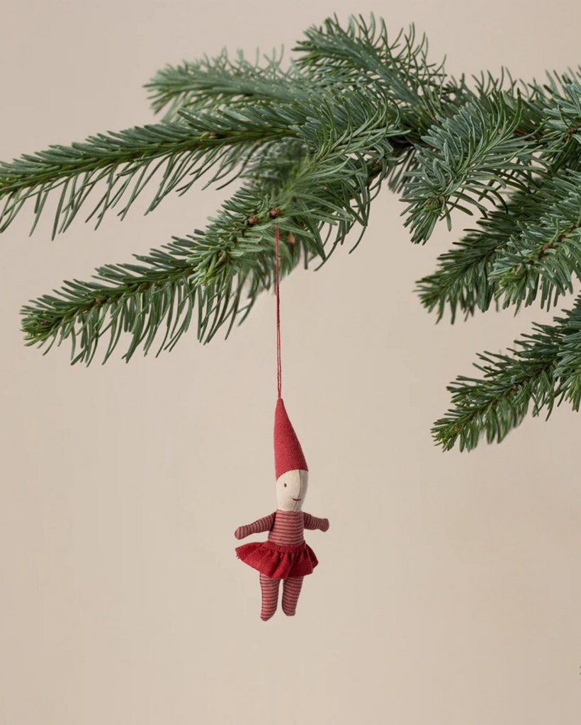 The Maileg Pixy Ornament - Girl, a small fabric doll with a red hat and striped outfit, hangs from a green pine branch, bringing festive charm to your Christmas decor against a neutral beige background.