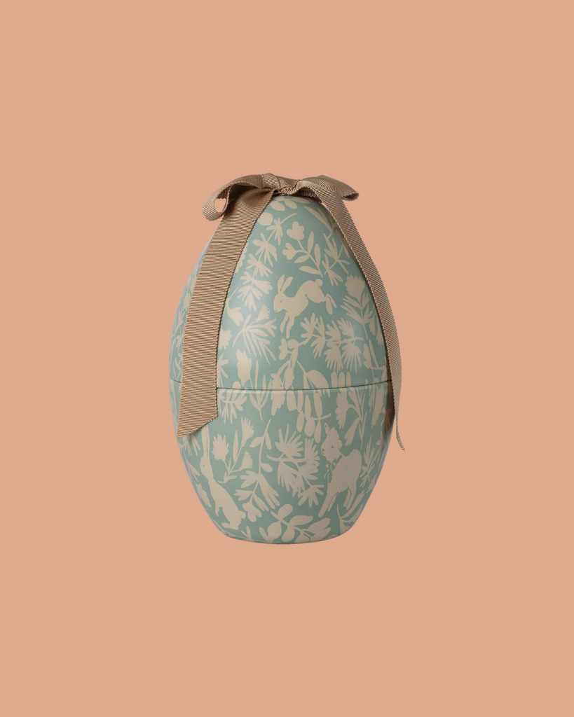 The Maileg Easter Egg, Medium - Dusty Blue, features a light blue and cream floral metal design with a beige ribbon bow, displayed against a peach background.