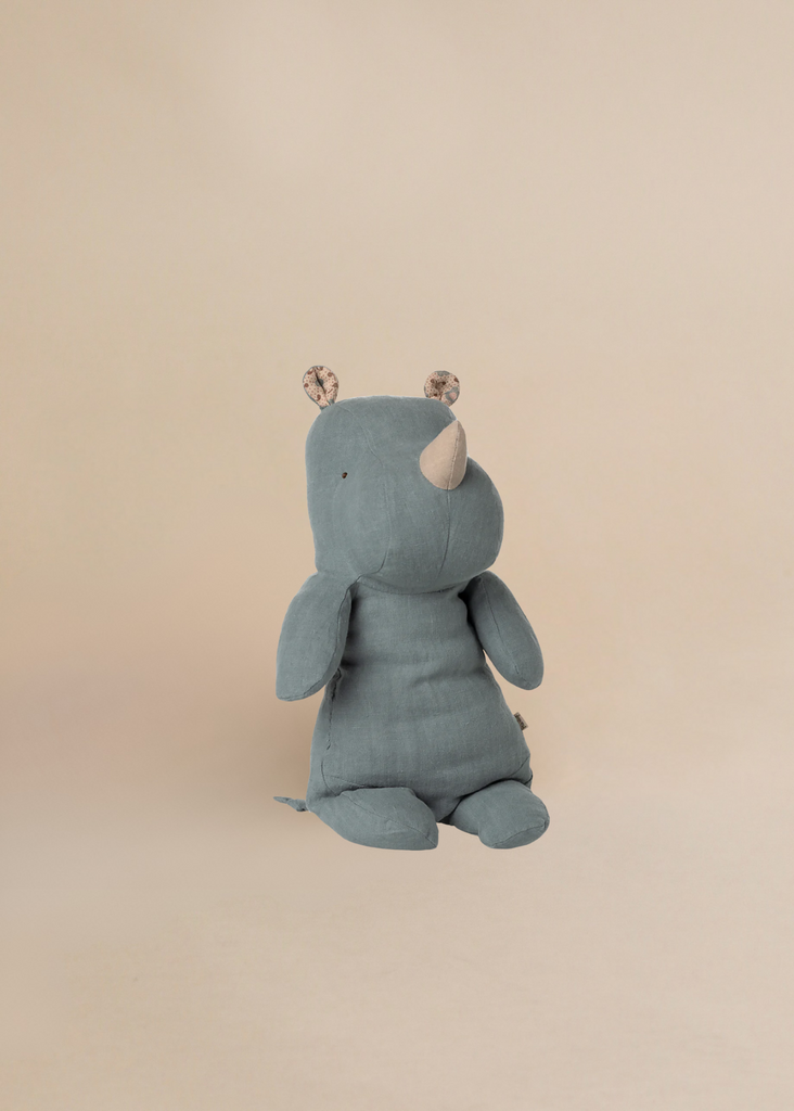 The Maileg Medium Rhino - Dusty Blue is a soft, blue-gray plush toy from our Safari Friends collection. Made from recycled polyester and soft fabric, it sits upright and features small patterned ears on a light beige background.