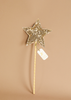 The Magic Wand - Gold Sequin Star features a sparkling gold sequin star, a wooden handle, and a small white tag, shown against a plain beige background.