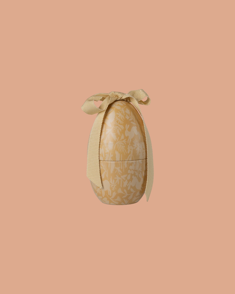 The Maileg Easter Egg, Small - Cream Yellow features a subtle floral pattern and a textured yellow ribbon at the top, shown against a solid peach background. Expected arrival: 02/15/26.