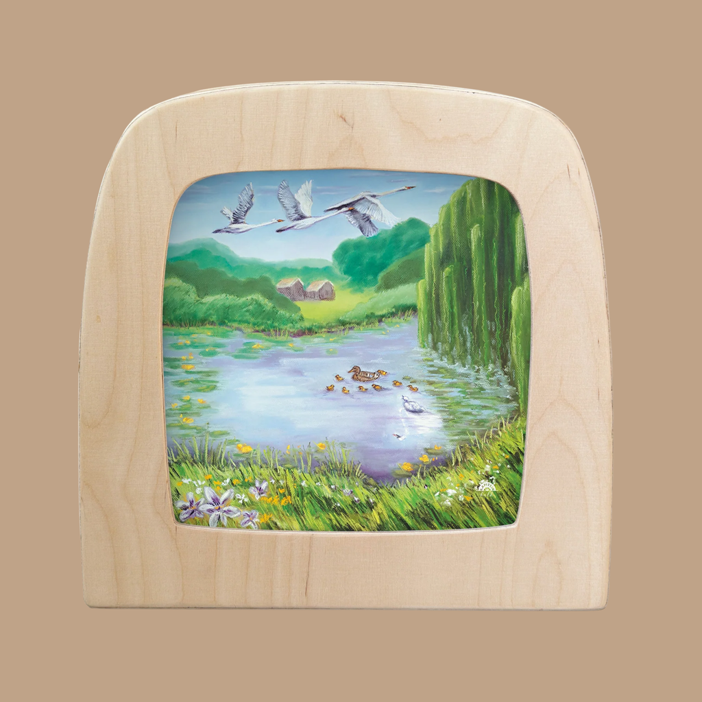 A wooden frame displays "Silhouette Only - The Ugly Duckling for Toverlux Lamp," featuring a pond with ducks, green trees, grass, flowers in front, and three cranes flying above two distant houses.