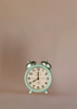 A small, vintage-style mint green alarm clock with silver bells is displayed alongside Maileg Grandma And Grandpa Afternoon Tea against a plain, light beige background.