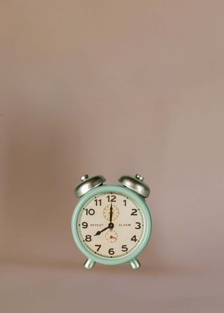 A small, vintage-style mint green alarm clock with silver bells is displayed alongside Maileg Grandma And Grandpa Afternoon Tea against a plain, light beige background.