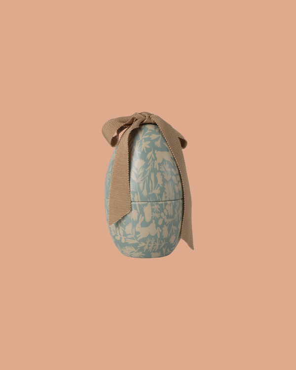 The Maileg Easter Egg, Small - Dusty Blue, features a floral pattern and a brown fabric ribbon tied in a bow on top, set against a peach-colored background. Expected arrival: 02/15/26.