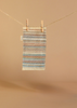 A small rectangular woven rug with horizontal multicolored stripes hangs on a twine clothesline using two wooden clothespins against a beige background—perfect for pairing with the Maileg Little Chef's Kitchen Set.
