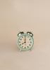 A small, vintage-style light blue alarm clock with silver bells sits against a plain beige background, making a charming addition to the Maileg Bedroom Set. The clock shows the time as 10:10.