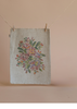 A piece of beige fabric with a colorful floral design, like the charming textiles in the Maileg Big Brother and Sister Afternoon Tea Set, hangs on a string with two wooden clothespins against a plain beige background.