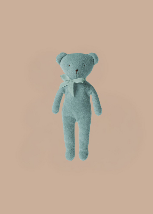 The Maileg Teddy - Blue is a classic light blue teddy bear made from soft fabric and adorned with a matching ribbon bow, shown against a simple peach background.